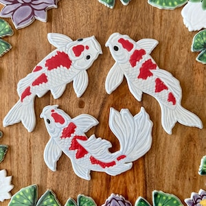 May include: Three white koi fish cookies with red markings and black eyes are arranged on a wooden surface. The cookies are surrounded by decorative floral and leaf-shaped cookies in shades of green, purple, and white.
