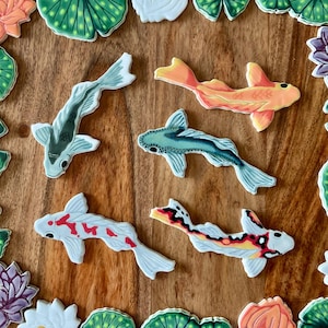 May include: Overhead shot of koi fish cookies in various colors, including orange, white, black, and gray. The cookies are arranged on a wooden surface, surrounded by lily pad and flower-shaped cookies.