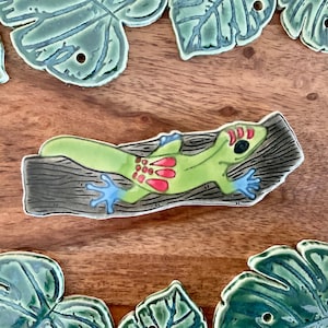 May include: A ceramic gecko lizard figurine with red and green accents, perched on a brown log. The figurine is painted with realistic details and has a glossy finish.