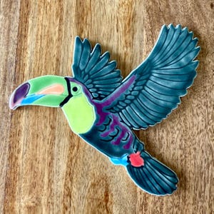 May include: A ceramic toucan bird figurine in flight, featuring a vibrant color palette. The bird has a large green and purple beak, a yellow chest, and blue wings with purple accents. The tail has red and blue details.