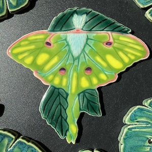 May include: A green and yellow ceramic tile with a detailed illustration of a luna moth. The moth has pink accents and is surrounded by green leaves.