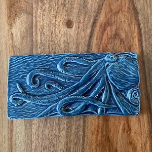 May include: A rectangular, blue ceramic tile featuring a detailed relief of an octopus. The octopus has multiple tentacles with suckers, and the tile has a glossy finish. The background has a wave-like texture.