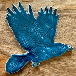 May include: A ceramic raven in flight, glazed in a vibrant teal blue. The bird's wings are outstretched, showcasing detailed feather patterns. The raven is set against a wooden surface.