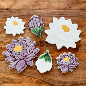 May include: Assortment of flower-shaped cookies with white, purple, and green icing. The cookies are decorated with yellow centers and sit on a wooden surface. The cookies are in the shape of lotus flowers and buds.