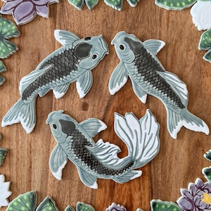 May include: Three ceramic koi fish in shades of gray and white are arranged on a wooden surface. The fish have detailed scales and fins, with a dark gray patch on their heads. Decorative lily pads and flowers surround the fish.
