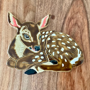May include: A ceramic fawn sculpture with brown and white spots, lying on its side with its head resting on its front paws. The fawn has pink ears and a light brown nose.