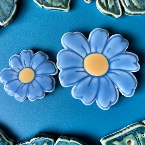 May include: Two ceramic flower tiles, one large and one small, with blue petals and a yellow center. The tiles are on a blue background.