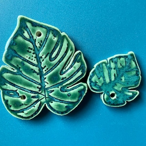 May include: Two ceramic monstera leaf ornaments in shades of green and blue. The larger leaf has a more detailed design and two holes. The smaller leaf has a simpler design and one hole.