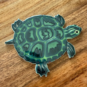 May include: A decorative turtle figurine with a green and dark green shell pattern. The turtle has a detailed design with visible legs, a head, and a tail. The figurine is placed on a wooden surface.