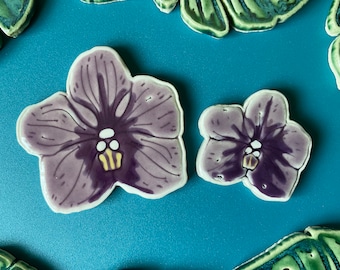 Könnte beinhalten: Zwei Keramikfliesen mit Orchideenblüten in Violett- und Gelbtönen. Die größere Fliese ist etwa 12,5 cm breit und die kleinere etwa 7,5 cm breit.