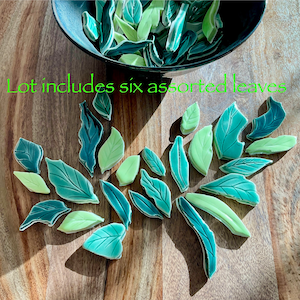 May include: A collection of ceramic leaf tiles in shades of green and teal, some inside a dark bowl, others scattered on a wooden surface. The text "Lot includes six assorted leaves" is visible. These decorative tiles are ideal for mosaic projects.