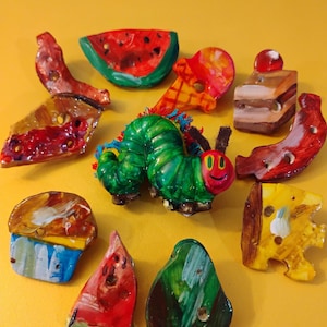 May include: A green caterpillar with a red smiling face surrounded by painted wooden food items, including a watermelon, a slice of cake, a banana, a cupcake, a pear, a slice of cheese, and an ice cream cone.