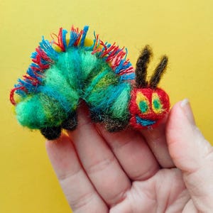 May include: A colorful, handmade felt caterpillar toy. The caterpillar features a red, yellow, and blue head with a smiling face and black antennae. The body is a mix of green, blue, and yellow, with red fringe. The toy is held against a yellow background.