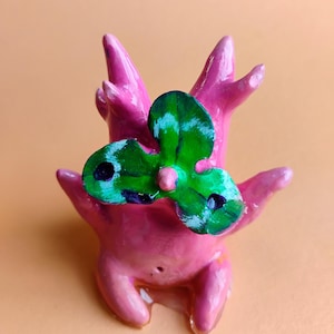 May include: A handmade pink sculpture of a creature with a green and white propeller-like design on its head. The sculpture has a smooth, glossy finish and is set against a peach-colored background.