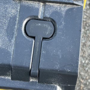 May include: Close-up of a black plastic latch on a yellow toolbox. The latch has a small, round handle and a long, thin lever.