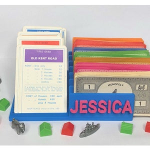 May include: A blue organiser with the name "JESSICA" in pink letters holds Monopoly game cards, title deeds, and play money. Game pieces include houses, hotels, and tokens.