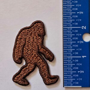 May include: Brown embroidered patch of a Bigfoot walking to the right. The patch is about 2 inches tall and 1.5 inches wide.