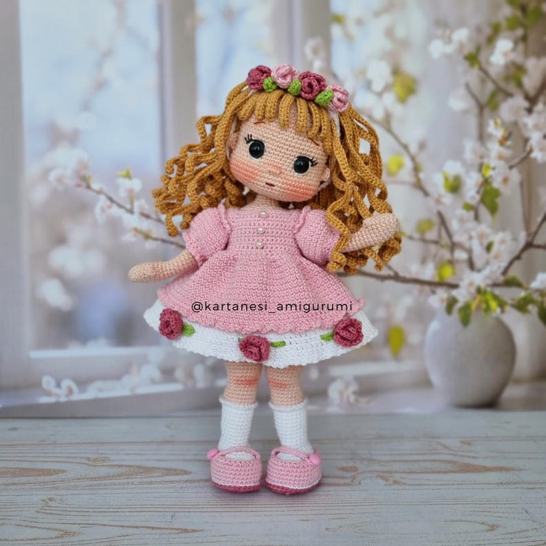Puede incluir: Una muñeca de crochet con el pelo rubio rizado, que lleva un vestido rosa con un ribete de encaje blanco y una corona de flores. La muñeca lleva zapatos rosas y calcetines blancos.