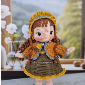 Puede incluir: Una mu&ntilde;eca de crochet con cabello casta&ntilde;o, vestida con un vestido amarillo y marr&oacute;n, una chaqueta azul y amarilla y un sombrero amarillo con ribete azul. La mu&ntilde;eca tiene una expresi&oacute;n dulce y est&aacute; extendiendo su brazo derecho.
