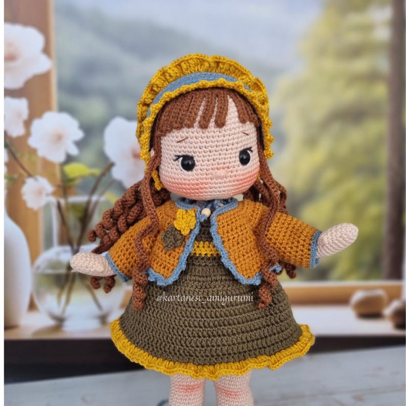 Puede incluir: Una mu&ntilde;eca de crochet con cabello casta&ntilde;o, vestida con un vestido amarillo y marr&oacute;n, una chaqueta azul y amarilla y un sombrero amarillo con ribete azul. La mu&ntilde;eca tiene una expresi&oacute;n dulce y est&aacute; extendiendo su brazo derecho.