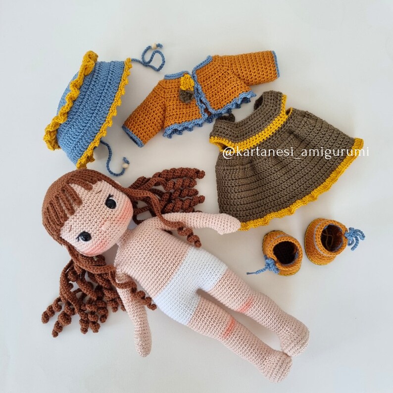 Puede incluir: Una mu&ntilde;eca de crochet con cabello marr&oacute;n, que lleva un pa&ntilde;al blanco y un vestido marr&oacute;n con ribetes amarillos. La mu&ntilde;eca tambi&eacute;n lleva un sombrero azul y amarillo y una chaqueta marr&oacute;n con ribetes amarillos. La mu&ntilde;eca est&aacute; acostada sobre una superficie blanca. @kartanes_amigurumi