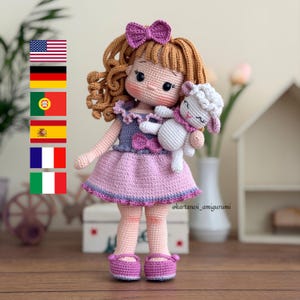 Puede incluir: Una muñeca de ganchillo hecha a mano con cabello castaño rizado, un lazo morado y un vestido rosa. La muñeca sostiene un pequeño cordero blanco. Los zapatos y el vestido de la muñeca están adornados con morado. La muñeca mide aproximadamente 25 cm de altura.