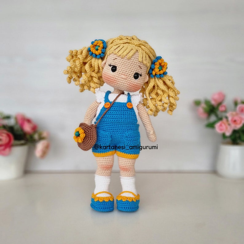 Puede incluir: Muñeca de ganchillo hecha a mano con cabello rubio rizado y ojos azules. La muñeca lleva un mono azul, una camiseta blanca y zapatos con ribete amarillo. Un pequeño bolso marrón con un detalle de girasol cuelga de su hombro.