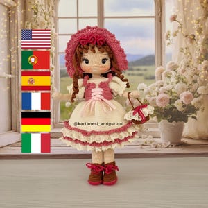 Puede incluir: Una muñeca de crochet que lleva un vestido rosa con ribete de encaje blanco, un sombrero rosa y zapatos marrones. La muñeca lleva una pequeña cesta de crochet blanca. La muñeca está de pie frente a una ventana con vista a un campo verde. El texto "@kartanes_amigurumi" es visible en el vestido de la muñeca.