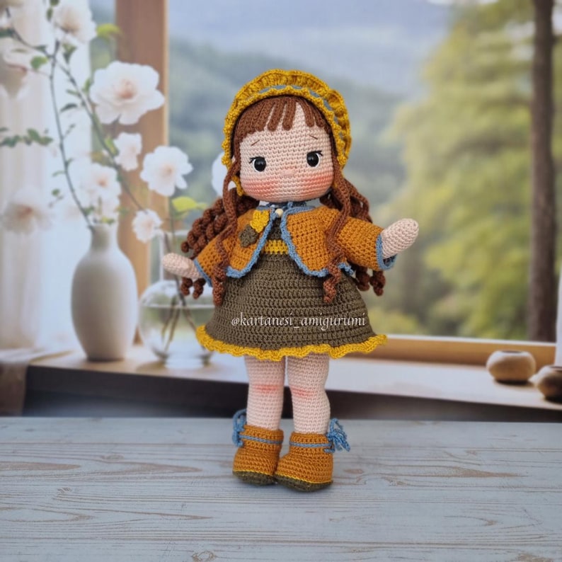 Puede incluir: Una mu&ntilde;eca de crochet con cabello casta&ntilde;o, vestida con un vestido amarillo y marr&oacute;n, una chaqueta marr&oacute;n y un sombrero amarillo con ribete azul. La mu&ntilde;eca est&aacute; de pie sobre una superficie de madera.