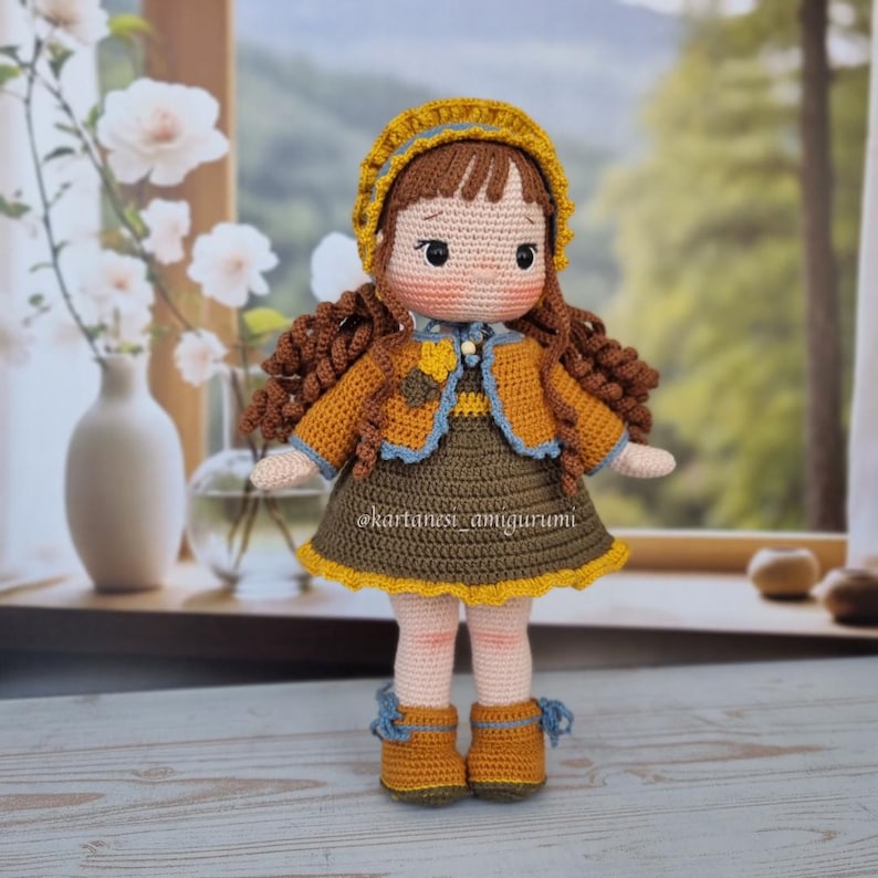 Puede incluir: Una mu&ntilde;eca de crochet con cabello casta&ntilde;o, que lleva un sombrero amarillo, una chaqueta marr&oacute;n, un vestido azul y marr&oacute;n y zapatos marrones. La mu&ntilde;eca est&aacute; de pie sobre una superficie de madera.