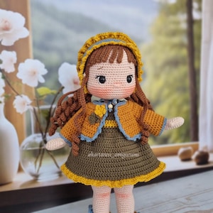 Puede incluir: Una mu&ntilde;eca de crochet con cabello casta&ntilde;o, vestida con un vestido amarillo y marr&oacute;n con ribete azul y un sombrero amarillo. La mu&ntilde;eca est&aacute; de pie sobre una superficie de madera.