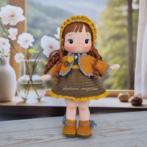 Puede incluir: Una mu&ntilde;eca de crochet con cabello casta&ntilde;o, que lleva un sombrero amarillo, un vestido marr&oacute;n con una chaqueta amarilla y zapatos marrones. La mu&ntilde;eca est&aacute; de pie sobre una superficie de madera.