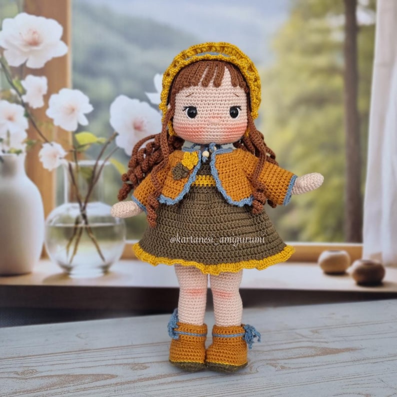 Puede incluir: Una mu&ntilde;eca de crochet con cabello casta&ntilde;o, que lleva un sombrero amarillo, un vestido marr&oacute;n con una chaqueta amarilla y zapatos marrones. La mu&ntilde;eca est&aacute; de pie sobre una superficie de madera.