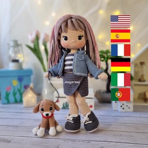 Puede incluir: Una muñeca de crochet con el pelo largo de color rosa que lleva una chaqueta vaquera azul, una camisa a rayas, una falda gris y zapatillas grises. La muñeca sostiene una correa atada a un pequeño perro de crochet marrón. La muñeca está de pie sobre una superficie de madera.