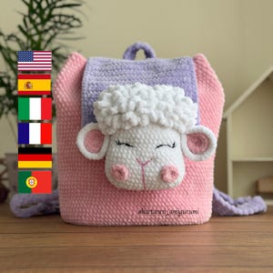 Könnte beinhalten: Häkelrucksack in Rosa und Lila mit einem weißen Schafgesicht. Das Schaf hat flauschige weiße Wolle, rosa Ohren und eine rosa Nase. Der Rucksack hat einen Griff und einen langen Riemen.