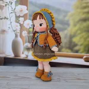 Puede incluir: Una mu&ntilde;eca de crochet con cabello casta&ntilde;o, que lleva un sombrero azul y amarillo, un vestido marr&oacute;n con ribete amarillo y zapatos marrones con detalles azules. La mu&ntilde;eca est&aacute; de pie sobre una superficie de madera.