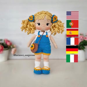 Könnte beinhalten: Handgemachte Häkelpuppe mit blonden Locken, die einen blauen Overall mit weißer Bordüre und eine braune Handtasche trägt. Die Puppe hat blaue Schuhe und ein Blumendetail an der Handtasche und im Haar. Die Puppe ist etwa 20 cm groß.