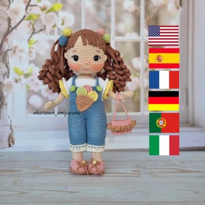 Puede incluir: Una muñeca de crochet con cabello castaño rizado, que lleva un vestido de mezclilla azul con un borde de encaje blanco, una camisa amarilla y blanca y zapatos rosas. La muñeca sostiene un bolso rosa de crochet. La muñeca está de pie sobre una superficie de madera.