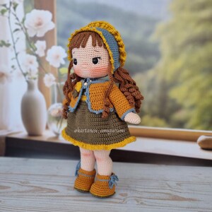 Puede incluir: Una mu&ntilde;eca de crochet con cabello casta&ntilde;o, vestida con un vestido amarillo y marr&oacute;n, un sombrero azul y amarillo y zapatos marrones. La mu&ntilde;eca est&aacute; de pie sobre una superficie de madera.