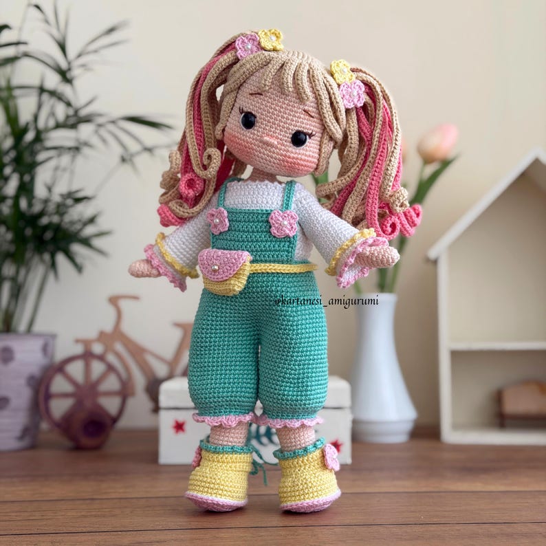Adorable crochet amigurumi doll with pink and yellow hair, floral outfit and belt bag; handmade soft toy for nursery decor and unique gift idea.