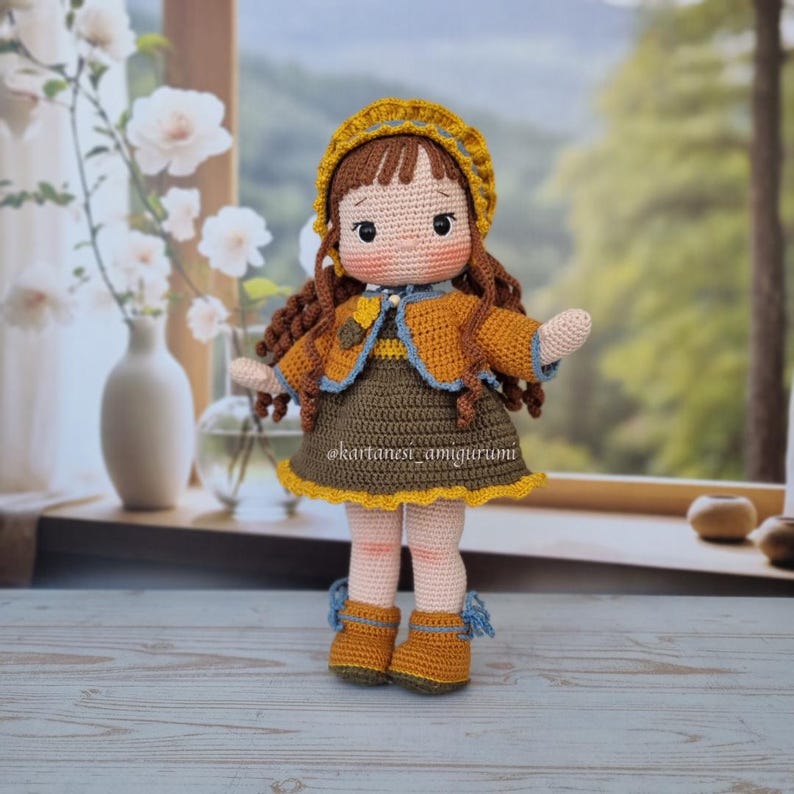 Puede incluir: Una mu&ntilde;eca de crochet con cabello casta&ntilde;o, vestida con un vestido amarillo y marr&oacute;n, una chaqueta marr&oacute;n y un sombrero amarillo con ribete azul. La mu&ntilde;eca est&aacute; de pie sobre una superficie de madera.