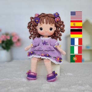 May include: A handmade crochet doll with brown curly hair, wearing a purple dress with flower details. The doll has a sweet expression and is wearing matching purple shoes. The text "@kartanesi_amigurumi" is visible on the dress.