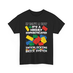 May include: Black t-shirt with a graphic featuring colorful interlocking toy bricks. The text reads "IT ISN'T A TOY IT'S A HIGHLY SOPHISTICATED INTERLOCKING BRICK SYSTEM". The design includes yellow, red, green, and blue bricks.