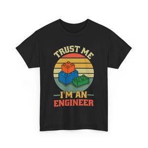 May include: Black t-shirt with a graphic of toy building blocks in orange, blue, and green, set against a retro sunset design. The text reads "TRUST ME I'M AN ENGINEER" in a vintage font.