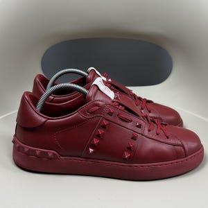 May include: A pair of red leather trainers with a white rubber sole and a studded design. The trainers have a lace-up closure and a low top.