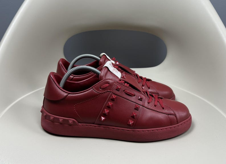 May include: A pair of red leather trainers with a white rubber sole and a studded design. The trainers have a lace-up closure and a low top.