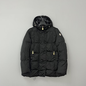 Puede incluir: Chaqueta acolchada negra con capucha, con herrajes dorados en los tiradores de la cremallera y los botones. La chaqueta tiene un diseño acolchado y un cordón en la parte inferior. La chaqueta está colgada en una percha sobre un fondo gris.