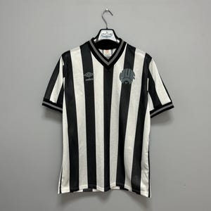 May include: Black and white striped soccer jersey with a V-neck collar and short sleeves. The jersey features the Umbro logo on the chest and a team emblem. The jersey is hanging on a hanger.