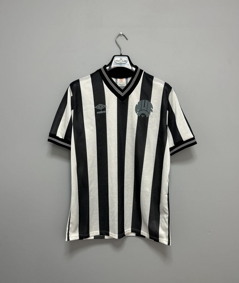 May include: Black and white striped soccer jersey with a V-neck collar and short sleeves. The jersey features the Umbro logo on the chest and a team emblem. The jersey is hanging on a hanger.