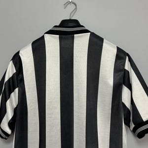 May include: Black and white striped jersey hanging on a metal hanger. The jersey has a black collar with white stripes and short sleeves. The stripes are vertical and evenly spaced.